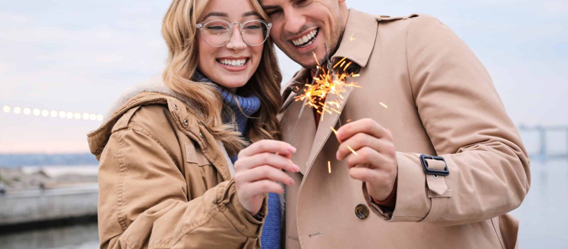 Couple in warm clothes holding burning sparklers near river